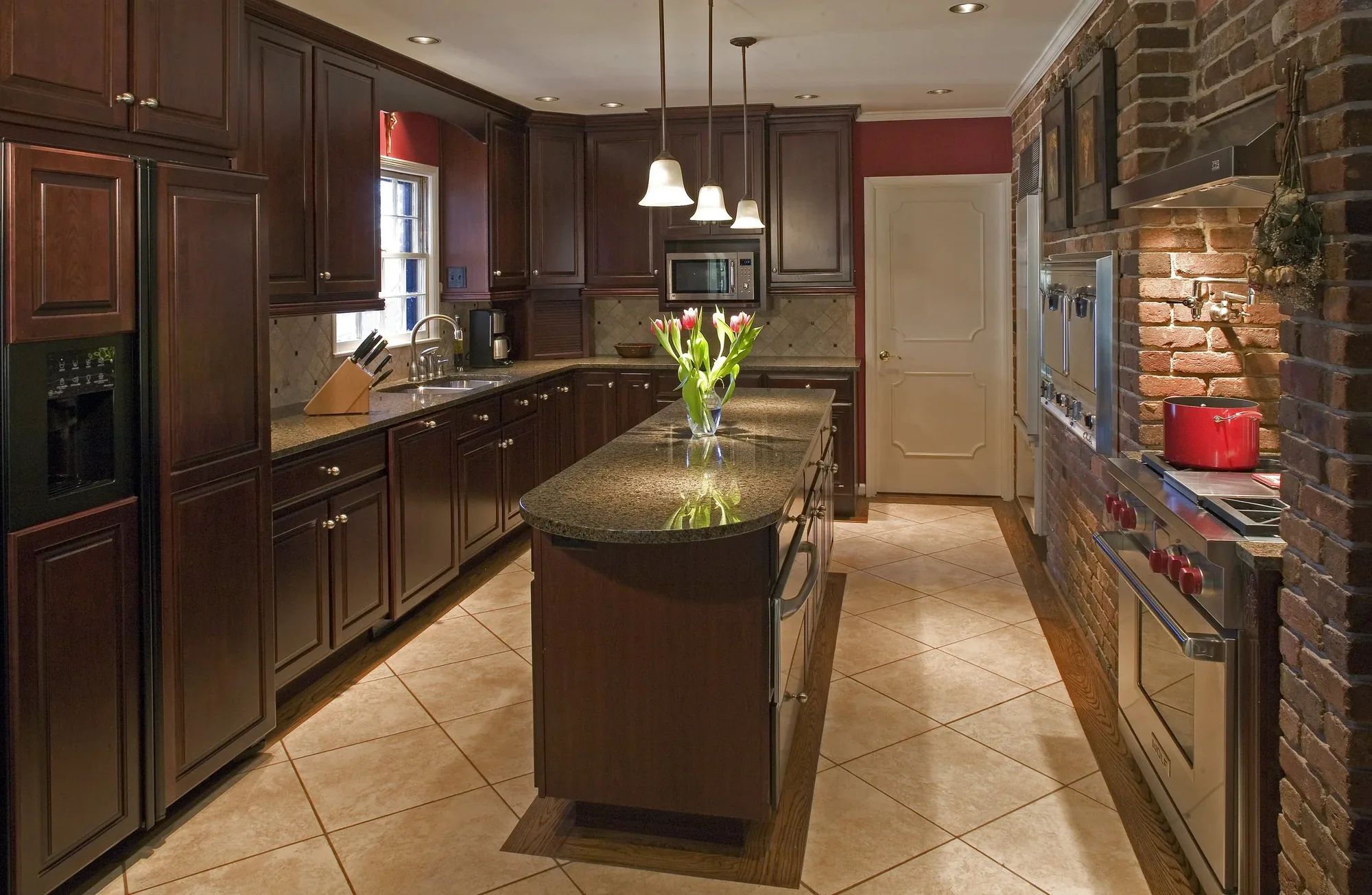 A long galley kitchen with dark wood cabinetry, a stone island and a brick wall.